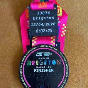 May include: A Brighton Marathon finisher medal with a black circular pendant featuring the text Brighton Marathon Finisher and 12 April 2026. A black rectangular tag above displays 33874, Brighton, 12/04/2026, and 6:02:25 on a bright pink ribbon.