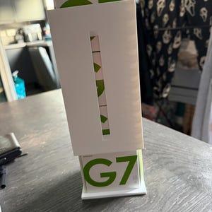 May include: A white display stand holding a green and white box labelled "G7". The stand has a rectangular cutout, revealing green and white elements. The product is on a dark grey wooden surface.