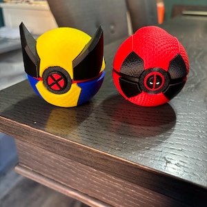 May include: Two spherical, 3D-printed objects. One is yellow, blue, and black, with a red X emblem. The other is red and black, with a Deadpool emblem. Both have a textured surface and are on a dark wooden table.