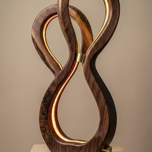 May include: A modern wooden sculpture with a warm, glowing interior light. The dark brown wood is shaped into intertwined loops, connected by a brass accent. The sculpture sits on a light-colored wooden block, creating a striking contrast.