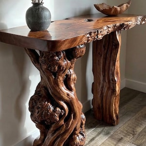 Op de afbeelding: Een handgemaakte houten console met een blad met levende rand en een unieke, organische basis. De tafel is gemaakt van rijk, donkerbruin hout en heeft een natuurlijke, getextureerde afwerking. Een donkergrijze vaas met gedroogde bloemen en een houten kom staan op de tafel.