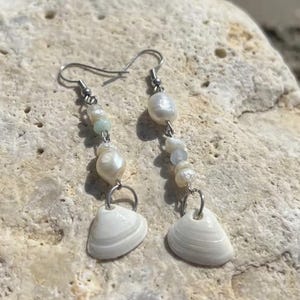 Könnte beinhalten: Hängeohrringe mit weißen Muscheln und Süßwasserperlen. Die Ohrringe haben silberfarbene Haken und bestehen aus kleinen, unregelmäßigen Perlen und einer einzelnen, größeren Muschel am unteren Ende. Der Hintergrund ist ein hellfarbiger Stein.