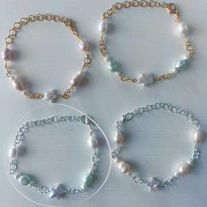 Op de afbeelding: Vier armbanden met zoetwaterparels en sterbedels. Twee armbanden hebben goudkleurige schakels en de andere twee hebben zilverkleurige schakels. De parels zijn wit, roze en lichtgroen.