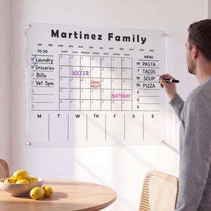 May include: A clear acrylic wall-mounted family calendar board featuring the text Martinez Family, a monthly grid, a to-do list, and a weekly menu section. The board includes printed black lines and headers for months, days, and tasks, mounted with silver hardware.