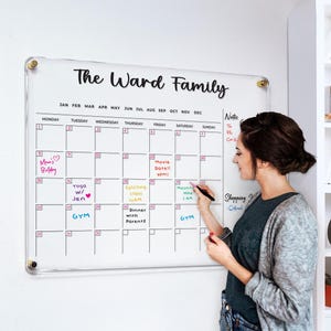 May include: A clear acrylic family calendar with the text "The Ward Family" at the top. The calendar grid is filled with handwritten notes and appointments. A woman is writing on the calendar with a black marker.