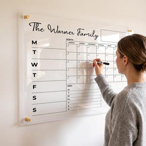 Może przedstawiać: Ścienny kalendarz wykonany z przezroczystego akrylu z czarnym nadrukiem The Warner Family, zawierający harmonogram tygodniowy, siatkę miesięczną oraz sekcję menu. Tablica jest przymocowana do ściany za pomocą czterech złotych metalowych dystansów.