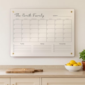 May include: A clear acrylic wall calendar featuring the text The Smith Family at the top, a monthly grid with days of the week, and three bottom sections labelled To Do, Notes, and Groceries, secured to the wall with four gold-toned metal standoffs.