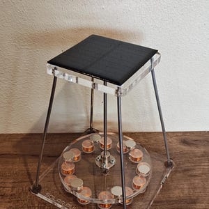 Peut inclure: Un moteur à impulsions solaires avec un panneau solaire noir sur le dessus, soutenu par des pieds en acrylique transparent. La base est en acrylique transparent avec le texte "SOLAR PULSE MOTOR". Le moteur a des bobines de cuivre.