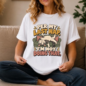 May include: A white short-sleeved t-shirt featuring a graphic of a cat sleeping on a green sofa. The text above the cat reads PER MY LAST NAP, and the text below the cat reads I'M NOT DOING THAT, in bold, multi-colored, rounded lettering.