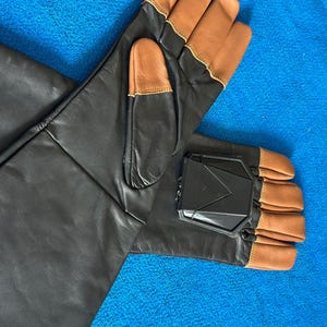 Pode incluir: Um par de luvas de couro preto com pontas e detalhes em couro castanho. As luvas t&ecirc;m uma pe&ccedil;a de pl&aacute;stico preto na parte de tr&aacute;s da m&atilde;o. As luvas est&atilde;o sobre uma superf&iacute;cie azul.