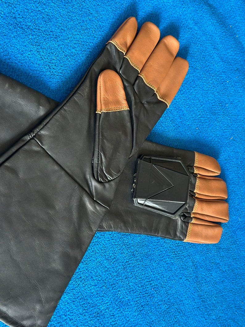 Pode incluir: Um par de luvas de couro preto com pontas e detalhes em couro castanho. As luvas t&ecirc;m uma pe&ccedil;a de pl&aacute;stico preto na parte de tr&aacute;s da m&atilde;o. As luvas est&atilde;o sobre uma superf&iacute;cie azul.