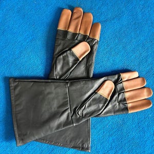 Pode incluir: Um par de luvas de couro preto com pontas e detalhes em castanho. As luvas est&atilde;o sobre uma superf&iacute;cie azul. As luvas parecem novas.