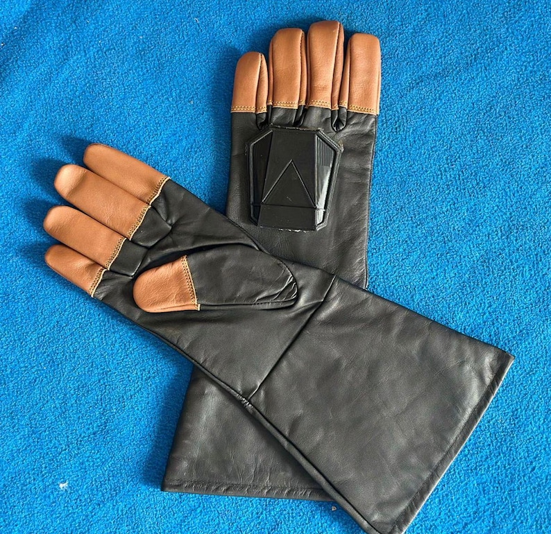 Pode incluir: Um par de luvas de couro preto com pontas dos dedos castanhas e um design geom&eacute;trico preto nas costas da m&atilde;o. As luvas t&ecirc;m punhos longos e est&atilde;o sobre uma superf&iacute;cie azul.