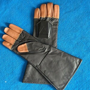 Pode incluir: Um par de luvas de couro preto com pontas dos dedos e detalhes na palma da m&atilde;o em castanho claro. Cada luva tem um design geom&eacute;trico preto nas costas da m&atilde;o. As luvas s&atilde;o exibidas em uma superf&iacute;cie azul.