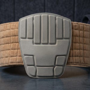 May include: A gray protective pad with a textured design, attached to a tan, quilted belt. The belt wraps around the pad, and the background is a dark gray.