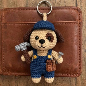 May include: Crocheted dog keyring wearing a blue cap and dungarees, holding a hammer and a wrench. The dog has a tan body with a brown spot and ears. The keyring is attached to a silver ring.