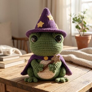 Puede incluir: Una rana de ganchillo hecha a mano que lleva un sombrero de mago morado con estrellas amarillas y una capa a juego. La rana es verde con grandes ojos negros y sostiene una varita mágica. La rana está sentada en una mesa de madera.