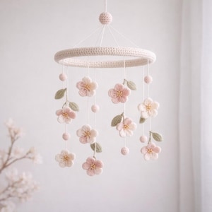 May include: A handcrafted crochet baby mobile with a circular frame and hanging flowers. The mobile features a cream-coloured ring with delicate pink and cream flowers, green leaves, and small pink spheres. The flowers have gold-coloured centres.