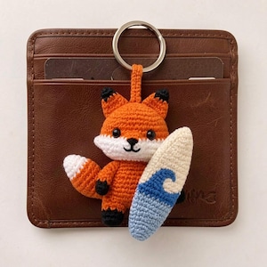 Puede incluir: Llavero de zorro tejido a ganchillo en naranja, blanco y negro, sosteniendo una pequeña tabla de surf con un diseño de ola azul. El llavero está sujeto a un anillo plateado y se coloca frente a una cartera de cuero marrón.