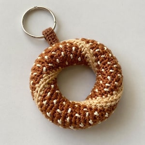 May include: A handmade crochet keychain in the shape of a bagel. The bagel is brown with white and tan accents, and a silver key ring is attached. The keychain is a unique accessory.