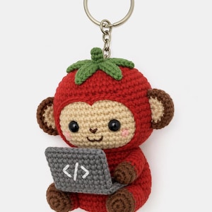 Peut inclure: Porte-clés crocheté représentant un singe déguisé en tomate, tenant un ordinateur portable gris avec des symboles de code. Le singe a des oreilles marron, un visage beige et des joues roses. Le porte-clés a un anneau argenté.