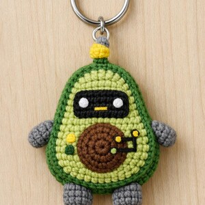 May include: Crocheted avocado-shaped keyring with a robot face. The green and yellow keyring has a brown seed, grey arms and legs, and a silver key ring. The robot face has a black rectangle with white eyes and a yellow line.