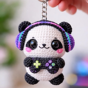Puede incluir: Llavero de panda tejido a crochet con temática de videojuegos. El panda tiene un cuerpo blanco, detalles negros y lleva auriculares con detalles morados, azules y rosas. Sostiene un mando negro con botones coloridos. Se adjunta un llavero plateado.