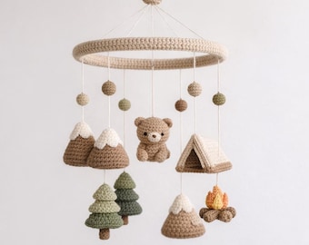 May include: A handmade crochet baby mobile featuring a circular frame and hanging elements. The mobile includes a bear, mountains, trees, a tent, and a campfire, all in neutral tones of brown, beige, and green. The mobile is suspended by a single string.