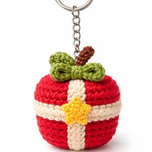 May include: A handmade crochet keychain in the shape of a red apple with a white stripe and a yellow star. The apple has a green bow and a brown stem. The keychain is attached to a silver metal ring.