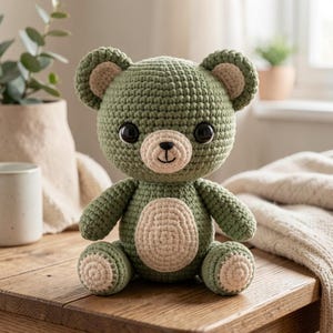May include: A sage green crocheted teddy bear with a cream-colored muzzle, belly patch, and inner ear accents. The stuffed toy features large, round, dark brown plastic eyes and a hand-stitched black nose and mouth, sitting in an upright position.