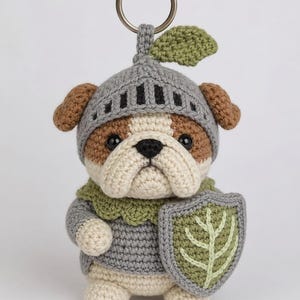 Puede incluir: Un llavero de bulldog tejido a ganchillo con un casco y túnica de caballero grises, sosteniendo un escudo con un diseño de hoja. El llavero tiene un anillo plateado y un acento de hoja verde en el casco. El bulldog tiene pelaje marrón y crema.