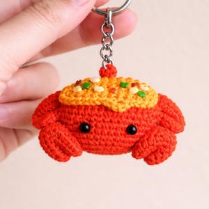 May include: A handmade crochet crab keychain in bright orange with a yellow and green topping. The crab has black eyes and is attached to a silver keyring. This whimsical accessory is perfect for anyone who loves marine life.