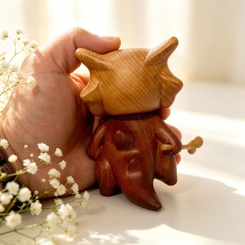 May include: A handcrafted wooden figurine with a light brown head and a dark brown body. The figurine has a unique, stylized design and is held in a person's hand. A small wooden stick is held in the figurine's hand. The background is blurred.