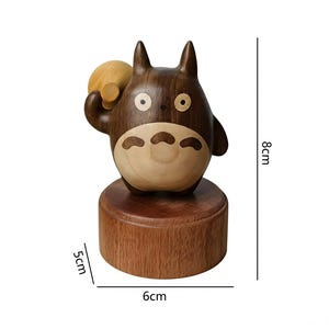 May include: A wooden Totoro figurine, 8cm tall, 6cm wide, and 5cm deep. The figure is brown with lighter wood accents on the belly and a small bag. The figurine is on a round wooden base.