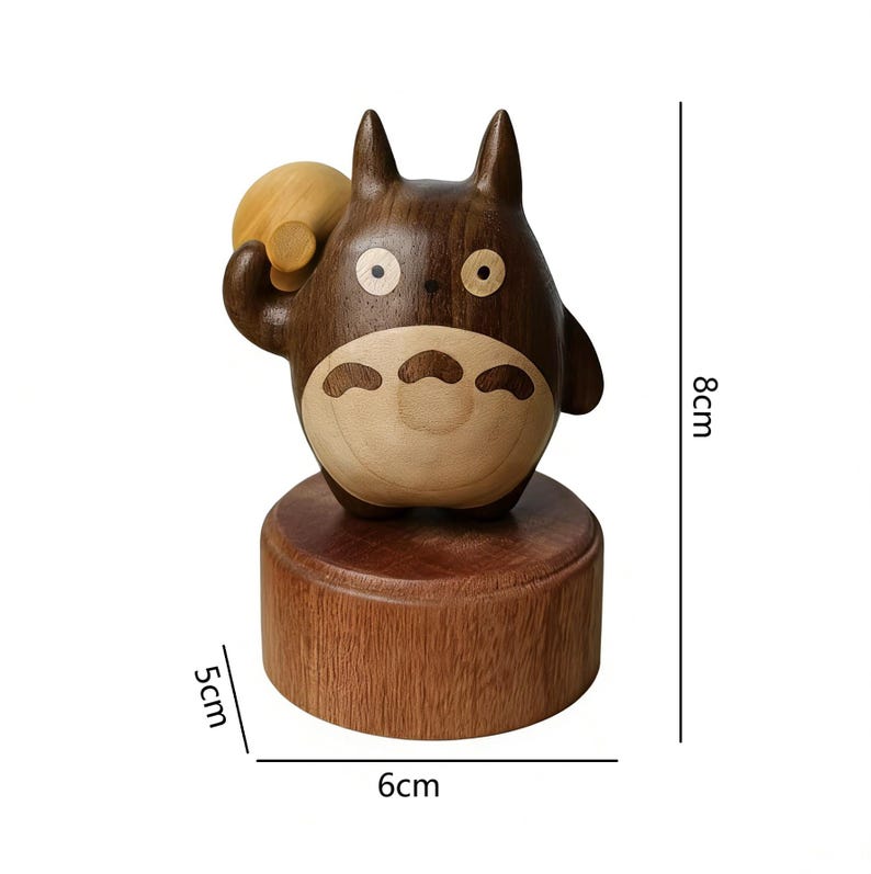 May include: A wooden Totoro figurine, 8cm tall, 6cm wide, and 5cm deep. The figure is brown with lighter wood accents on the belly and a small bag. The figurine is on a round wooden base.