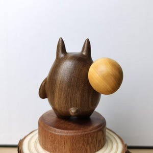 May include: A wooden figurine of a cartoon character with a round body, two pointed ears, and a small tail. The figurine is dark brown and holds a light brown ball. It sits on a wooden base atop a tree slice.