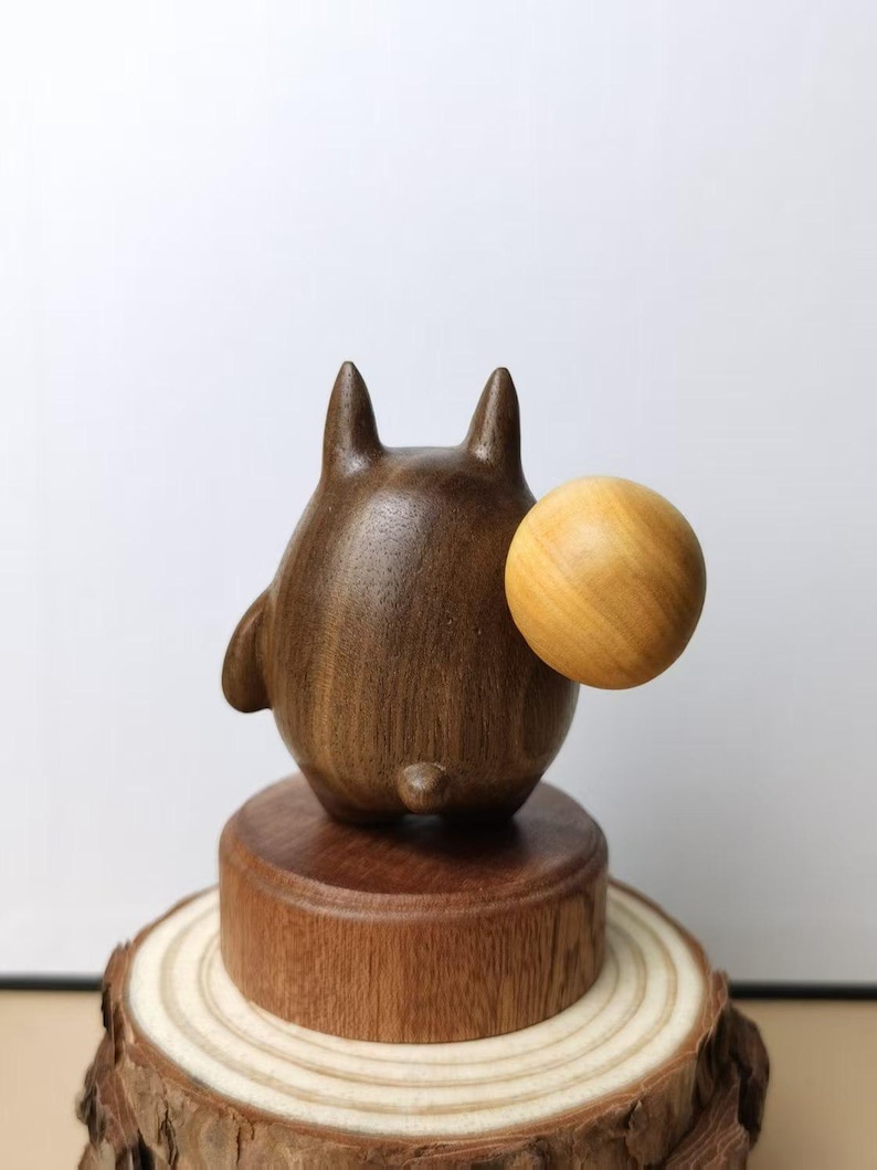 May include: A wooden figurine of a cartoon character with a round body, two pointed ears, and a small tail. The figurine is dark brown and holds a light brown ball. It sits on a wooden base atop a tree slice.
