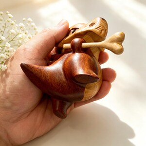 May include: A handcrafted wooden dinosaur figurine, holding a bone in its mouth. The dinosaur is made of two tones of wood, with a darker brown for the body and a lighter tan for the head and bone. The figurine is held in a hand.