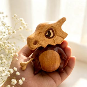 May include: A handcrafted wooden Cubone figure, featuring a light brown skull-like head and a dark brown body. The figure is held in a hand, showcasing the detailed wood grain and craftsmanship. The bone club is also made of wood.