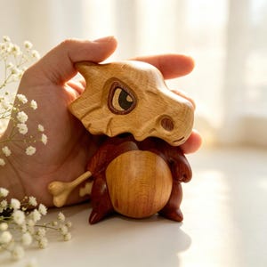 May include: A handcrafted wooden Cubone figure, featuring a light brown skull-like head and a dark brown body. The figure has a bone accessory and is held in a hand, showcasing the detailed wood grain and craftsmanship. The figure is a collectible item.