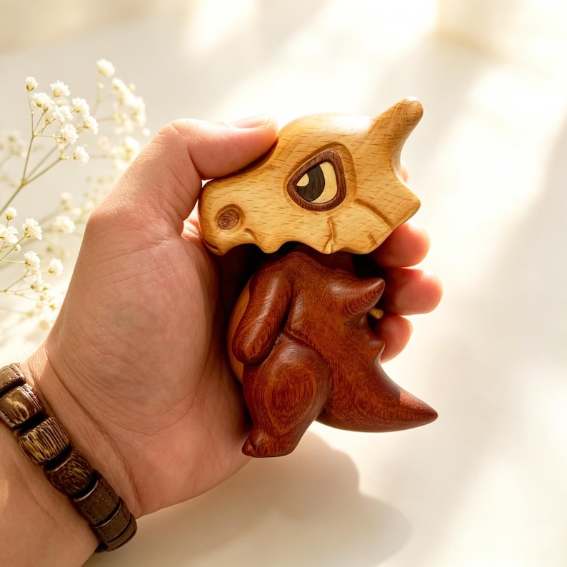 May include: A wooden Cubone figurine, featuring a light brown skull and a dark brown body. The figurine is held in a hand, showcasing the detailed craftsmanship and the contrast between the two wood tones. The eye is a dark brown color.