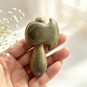 Può includere: Uno strumento per massaggio in legno a forma di piccola ascia. Lo strumento è di colore verde-marrone tenue con venature del legno visibili. L'impugnatura è a forma di goccia e la testa ha un design curvo. Lo strumento è tenuto in una mano.
