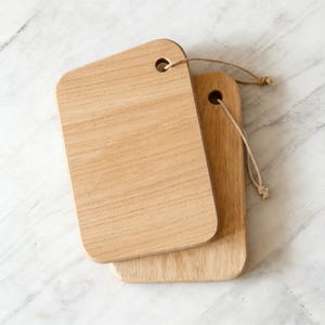 Puede incluir: Dos tablas de cortar rectangulares de madera con esquinas redondeadas y un agujero para colgar. Las tablas son de color marrón claro con vetas de madera visibles y están unidas por una cuerda natural. Las tablas están apiladas.