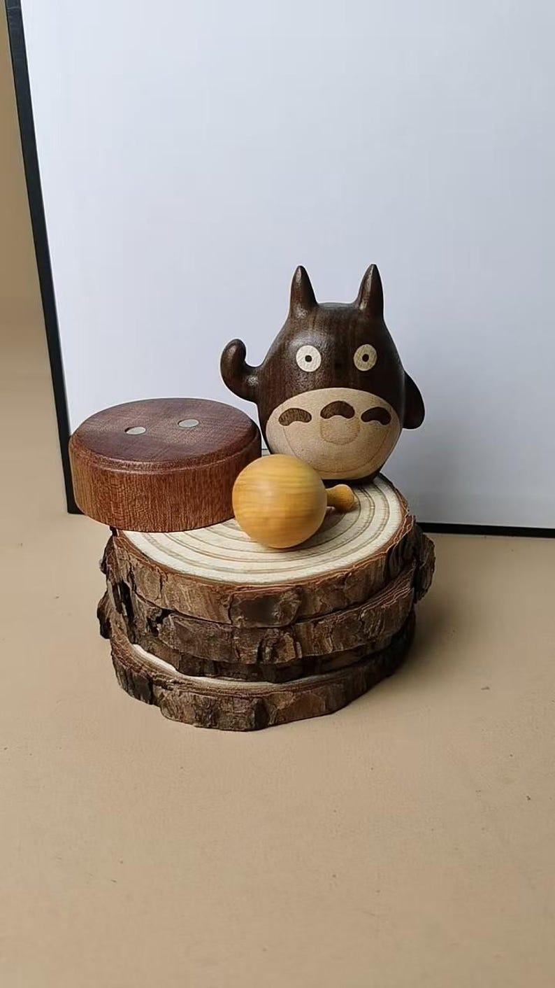 May include: A wooden Totoro figurine, a round wooden ball, and a wooden lid, displayed on a stack of wood slices. The Totoro is dark brown with a light brown belly and white eyes. The wood slices have a natural bark edge.