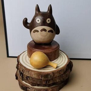 May include: A wooden Totoro figurine with a light brown belly and face, dark brown body, and round eyes. It sits atop a wooden base and a stack of wood slices. A small, wooden rattle rests on the wood slices.