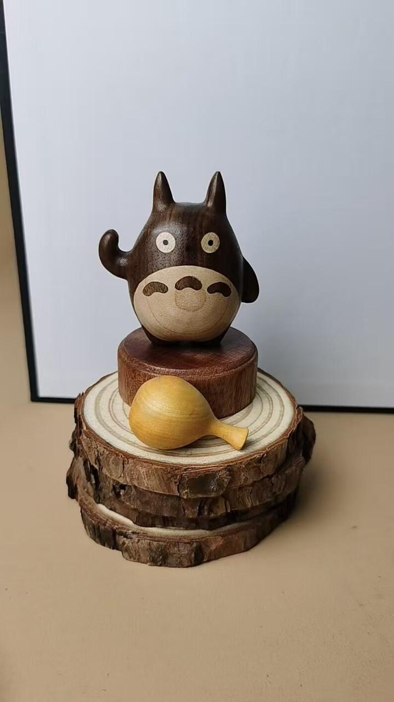 May include: A wooden Totoro figurine with a light brown belly and face, dark brown body, and round eyes. It sits atop a wooden base and a stack of wood slices. A small, wooden rattle rests on the wood slices.
