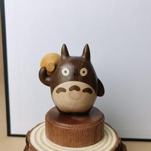 May include: A wooden figurine of Totoro, a character from the animated film My Neighbor Totoro. The figurine is brown and tan, with a round body and pointed ears. It is holding a small, round object and is mounted on a wooden base.
