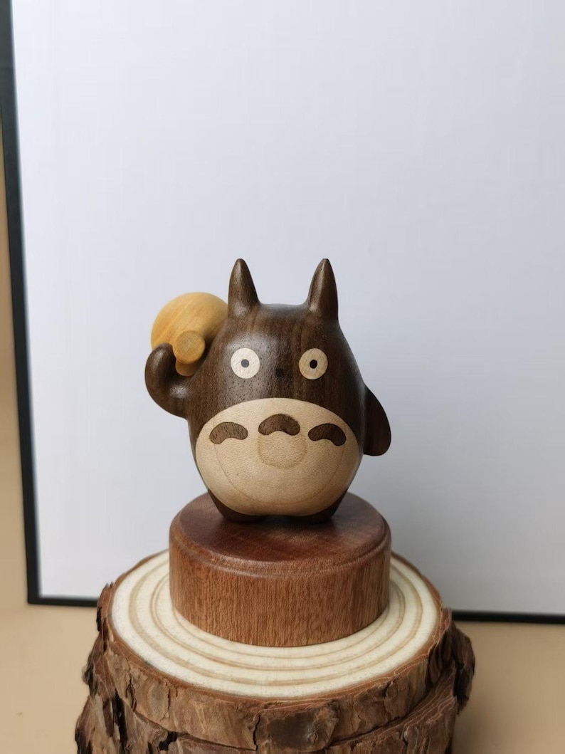 May include: A wooden figurine of Totoro, a character from the animated film My Neighbor Totoro. The figurine is brown and tan, with a round body and pointed ears. It is holding a small, round object and is mounted on a wooden base.