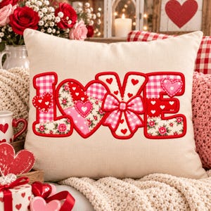 May include: A square off-white throw pillow featuring the word LOVE appliqued in red and pink patterns. The letters are decorated with gingham, floral, and heart motifs, with a pink gingham bow centered on the letter V and red stitching.