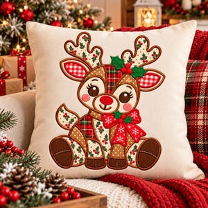 May include: A square off-white throw pillow featuring a central embroidered design of a cartoon reindeer. The reindeer has patchwork-style patterns in red, green, and brown, with holly leaves, berries, and a large red bow with white snowflakes.
